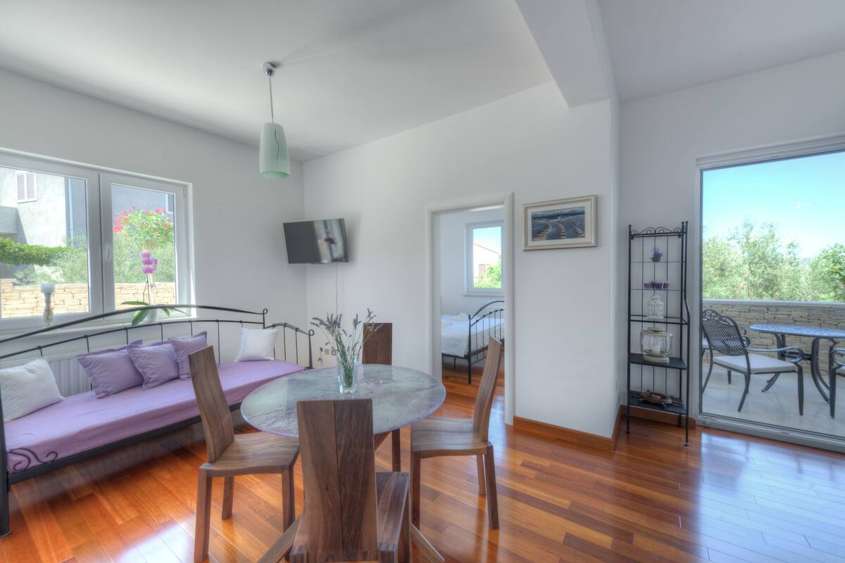A modern living room with a sofa, dining table, and chairs, a TV mounted on the wall, and a balcony with outdoor furniture.