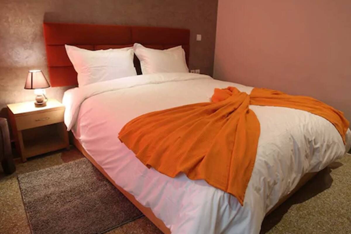 A neatly made bed with white and orange bedding, a wooden nightstand with a lamp, and a red headboard.