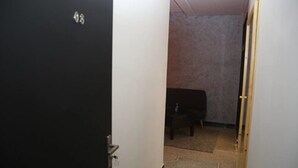 Een smalle gang met een deur waarop '48' staat, die leidt naar een kamer met een bed en een kleine tafel.