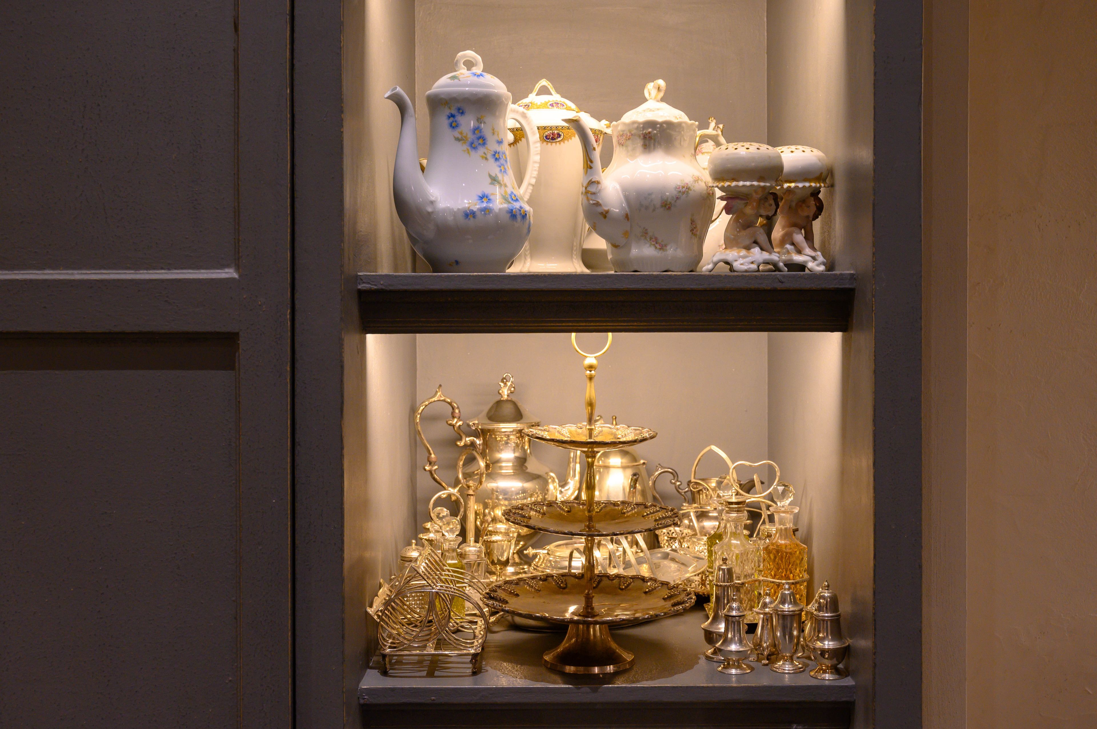 A display of antique tea sets and silverware on shelves.