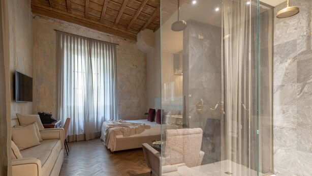 A modern bedroom with a wooden ceiling, a bed with a white headboard, a beige sofa, and a glass-enclosed shower area with marble walls. - Residenza La Musa Amarcord (Florence)