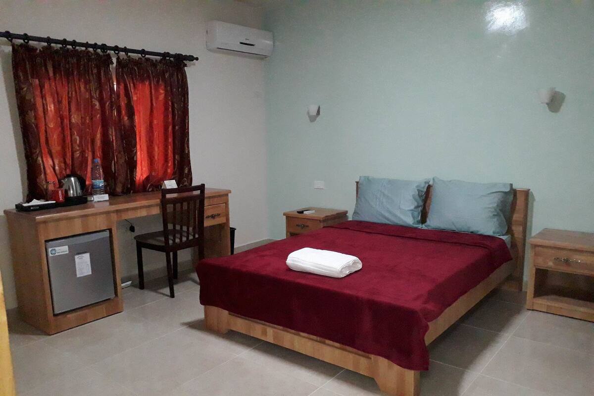 A room with a bed, a desk, a chair, a small refrigerator, and a nightstand.
