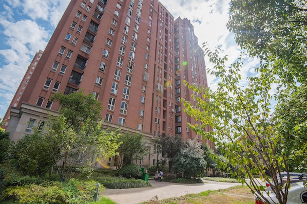 A tall red brick building with multiple floors and balconies, surrounded by greenery and a paved area.
