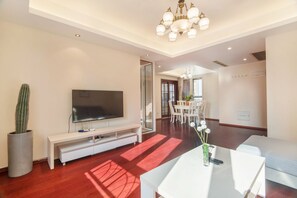 A modern living room with a flat-screen TV, a white sofa, a dining area with a table and chairs, and a large potted cactus. - Central Park (Suzhou)
