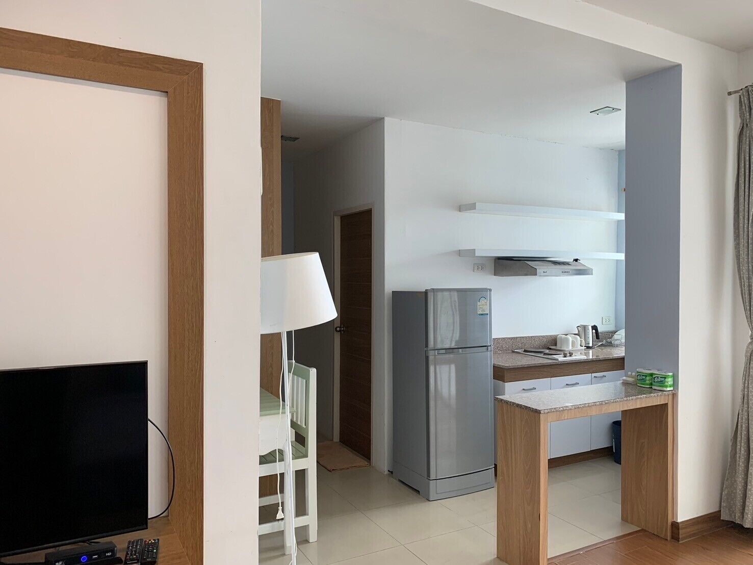 A compact hotel room with a TV, a desk with a lamp, a kitchenette with a fridge, and a shower area.