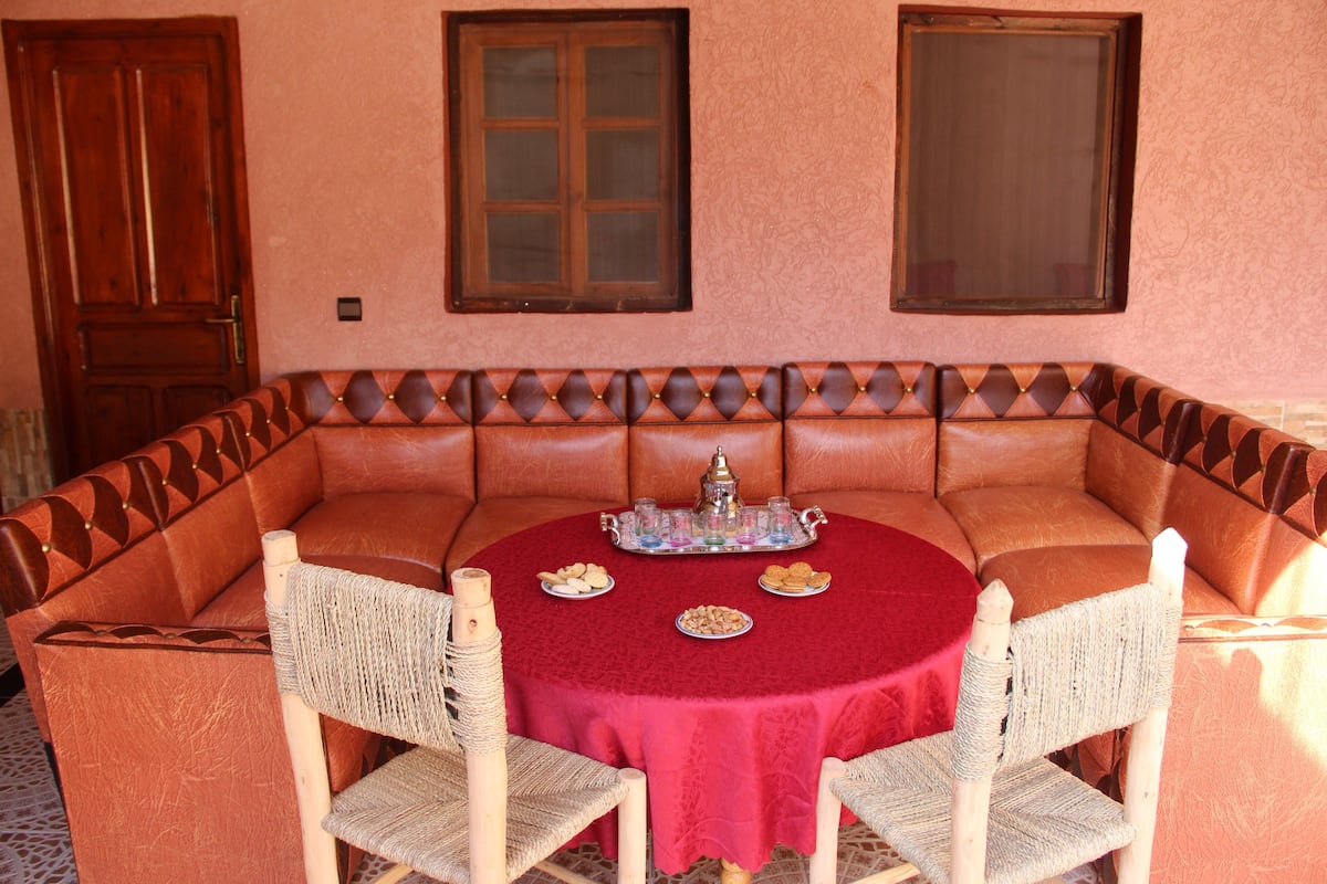 A round table with a red tablecloth, surrounded by wicker chairs, set against a peach-colored wall with two windows.