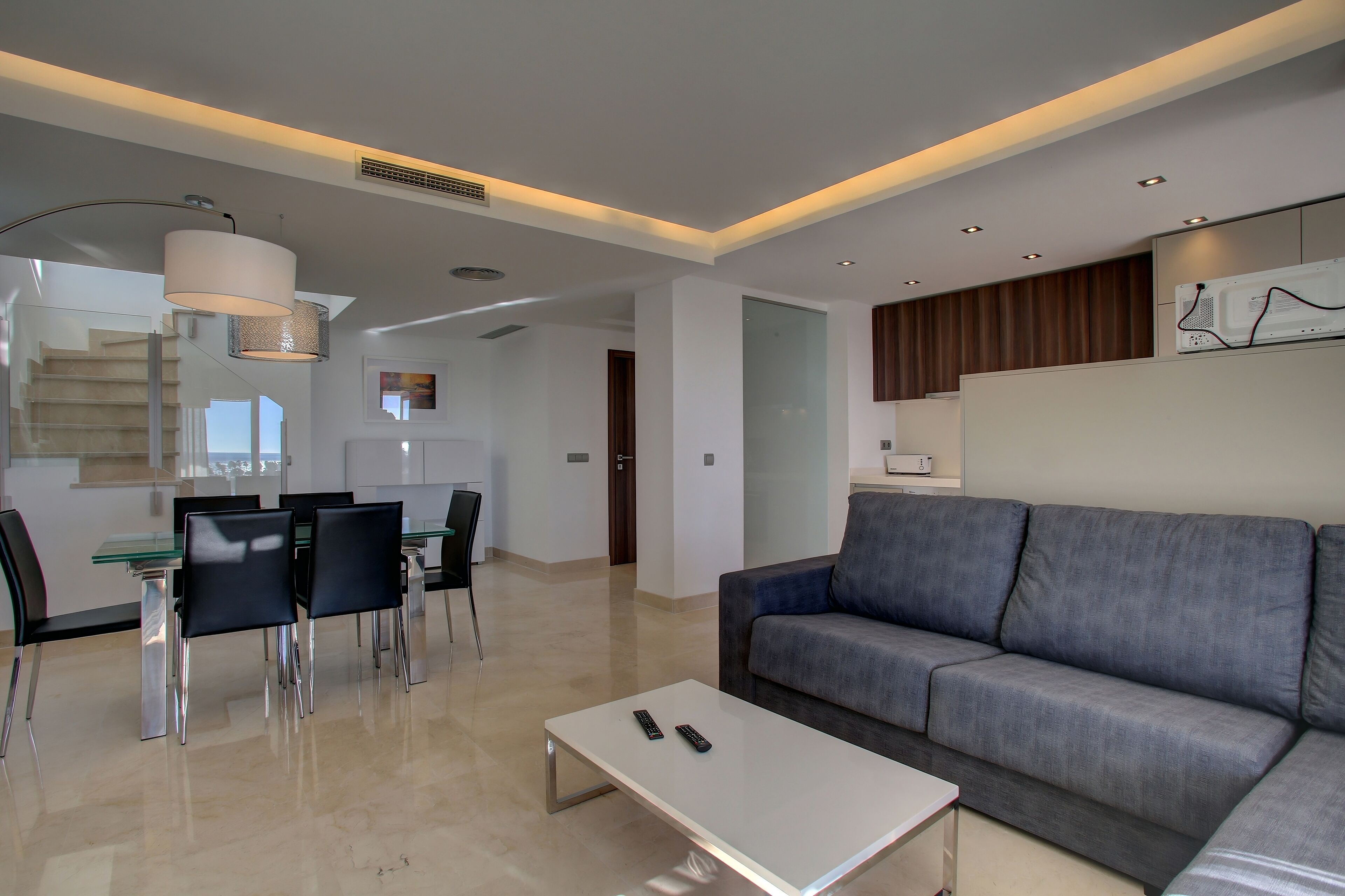 A modern living room with a grey sofa, a glass dining table with chairs, a white coffee table, and a staircase leading to an upper level.