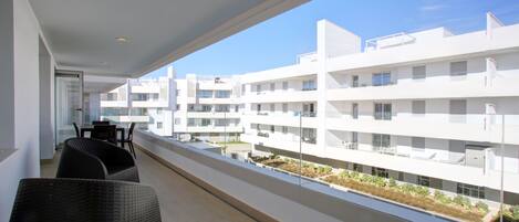 A balcony with wicker furniture, a clear view of a white building complex, and a well-lit interior.