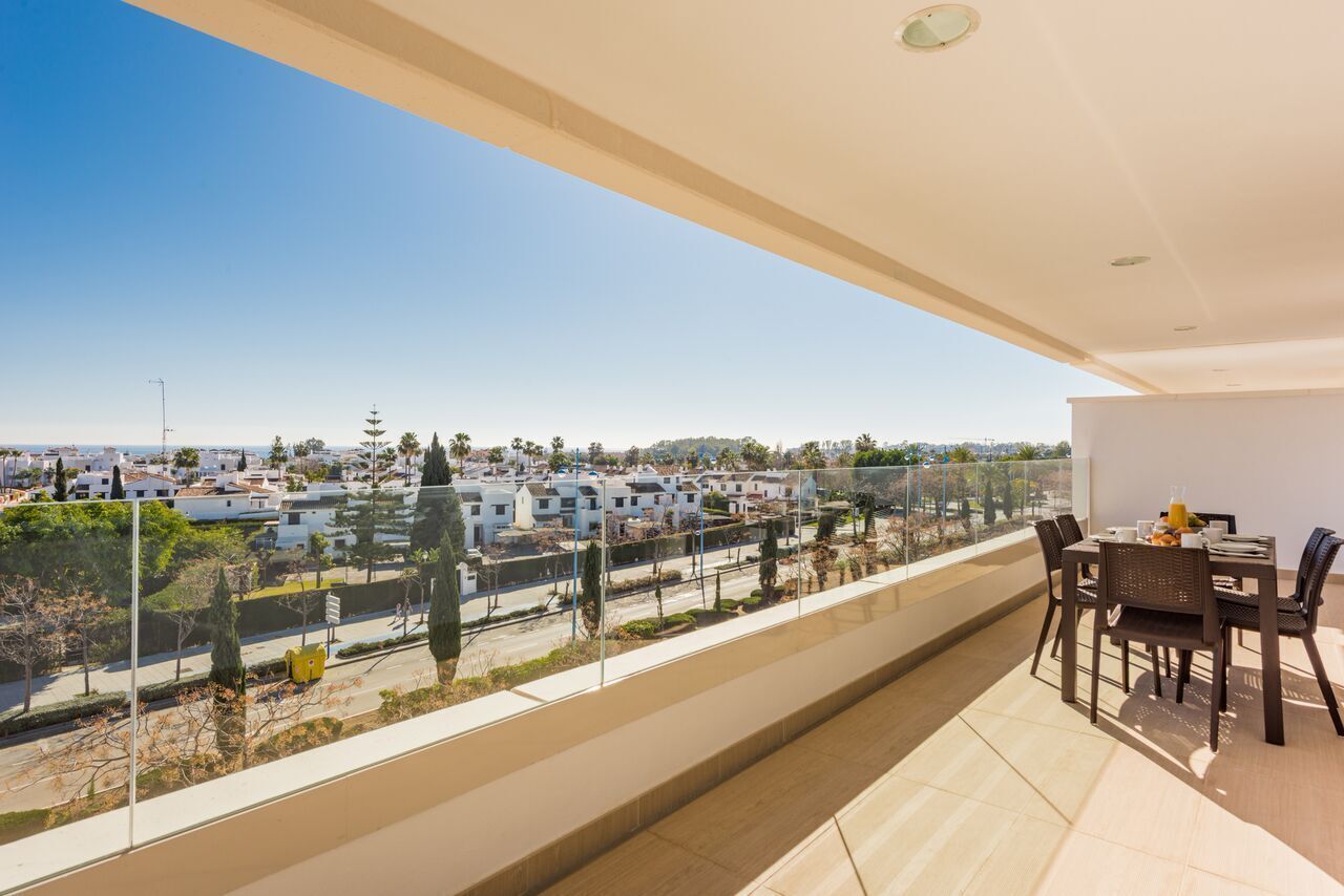 Un balcón con vista a una zona residencial, una mesa de comedor con sillas y cielo despejado.