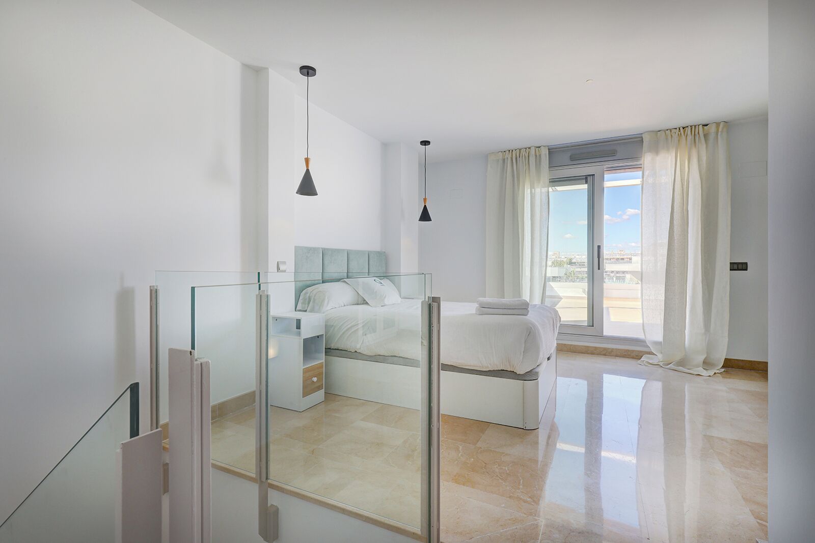 A modern bedroom with a glass partition, a bed with white bedding, a large window with curtains, and a city view.