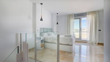 A modern bedroom with a glass partition, a bed with white bedding, a large window with curtains, and a city view.