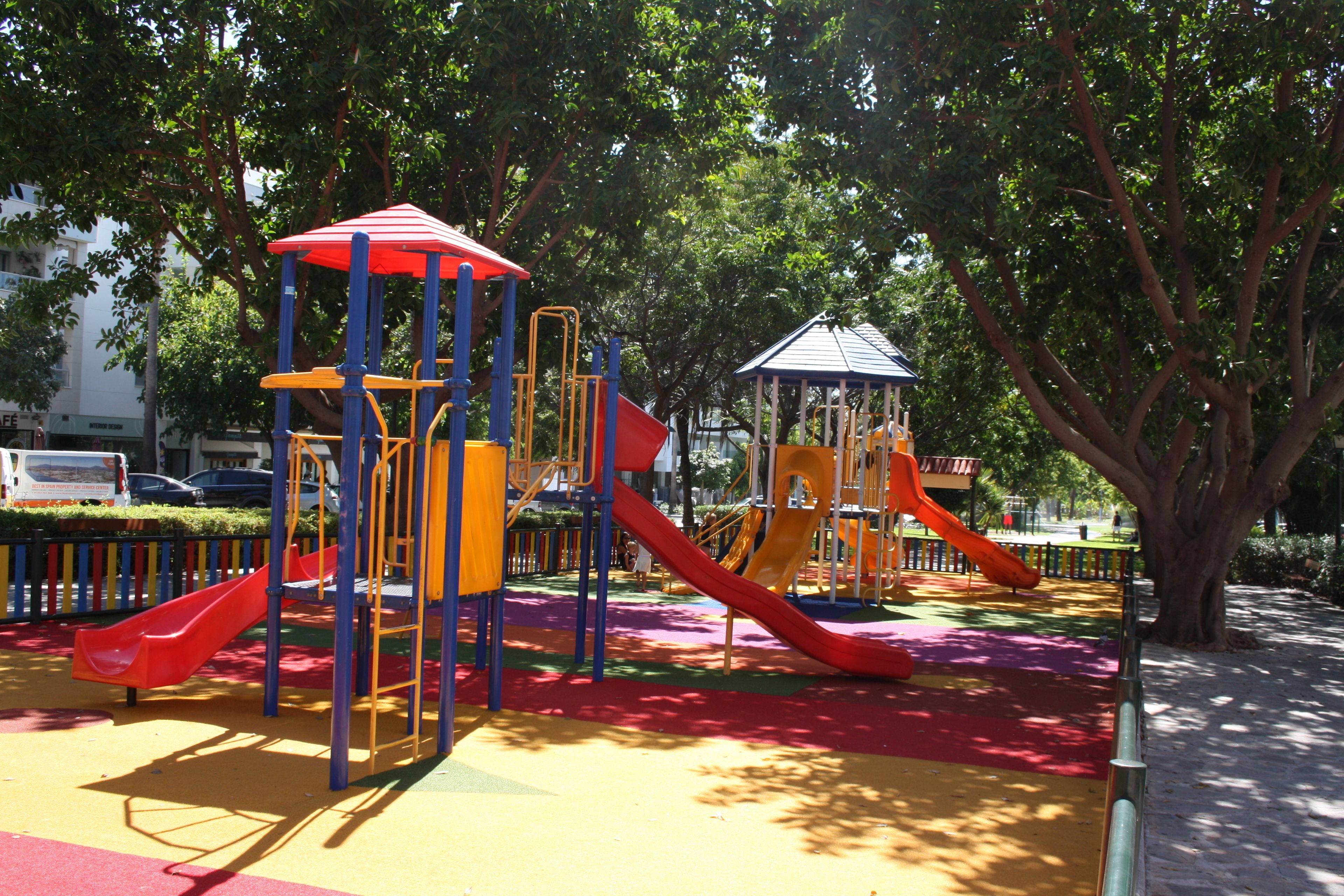 Zona de jocs infantil amb tobogans i elements per escalar, envoltada d'arbres i una superfície pavimentada.