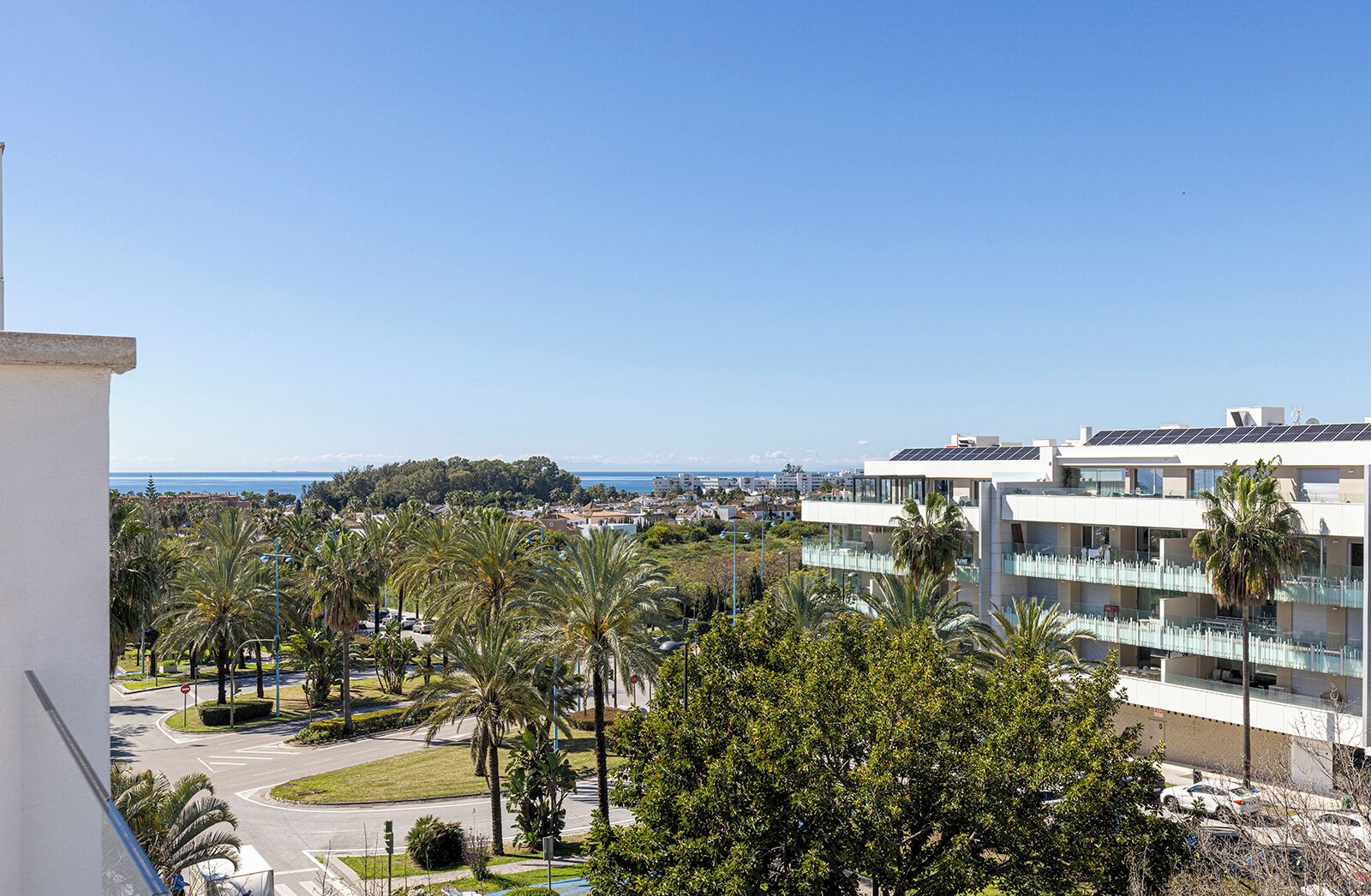 A modern hotel with multiple floors, a view of the sea, and a parking area with palm trees.