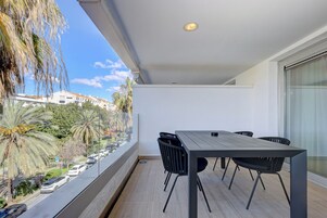 Een balkon met een tafel en stoelen, met uitzicht op een straat met geparkeerde auto's en palmbomen.