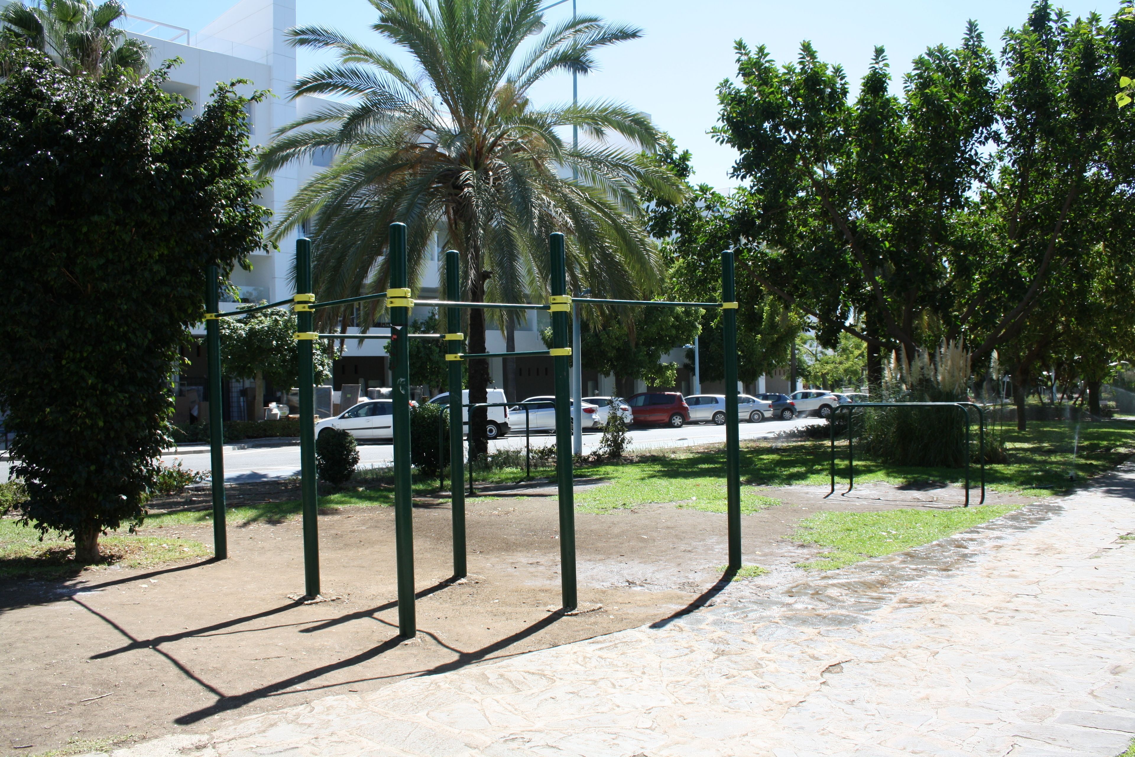 Un espacio exterior para ejercitarse con una estructura metálica, rodeado de árboles y una carretera con coches.