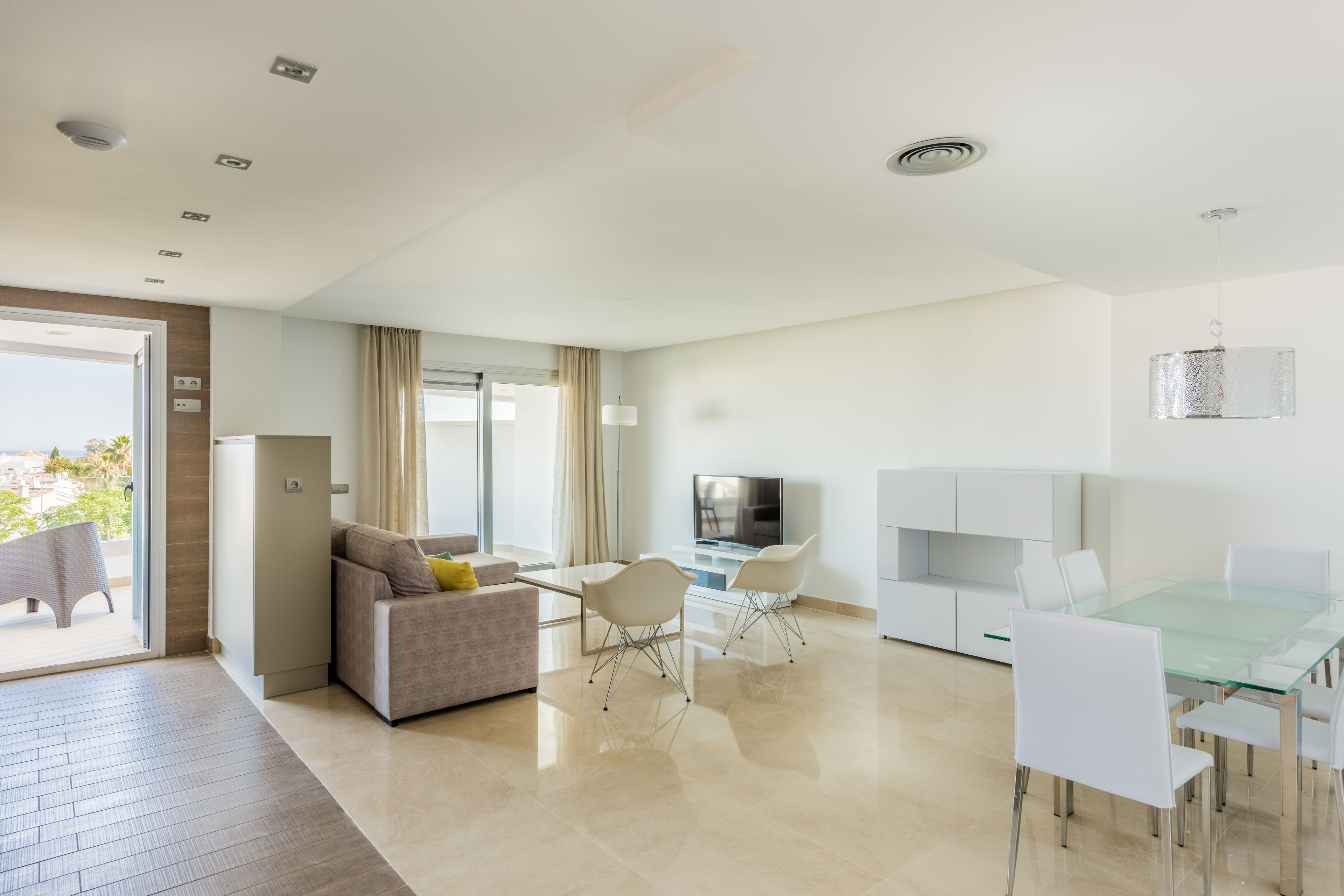 A modern living room with a sofa, a dining table with chairs, a TV, and a balcony with a view.