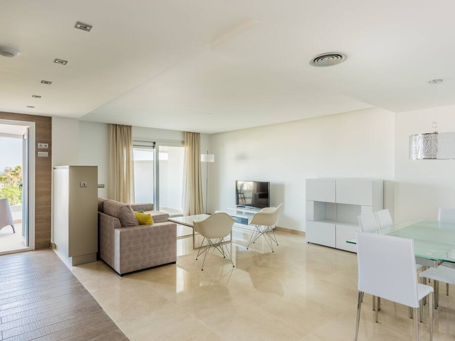 A modern living room with a sofa, a dining table with chairs, a TV, and a balcony with a view.