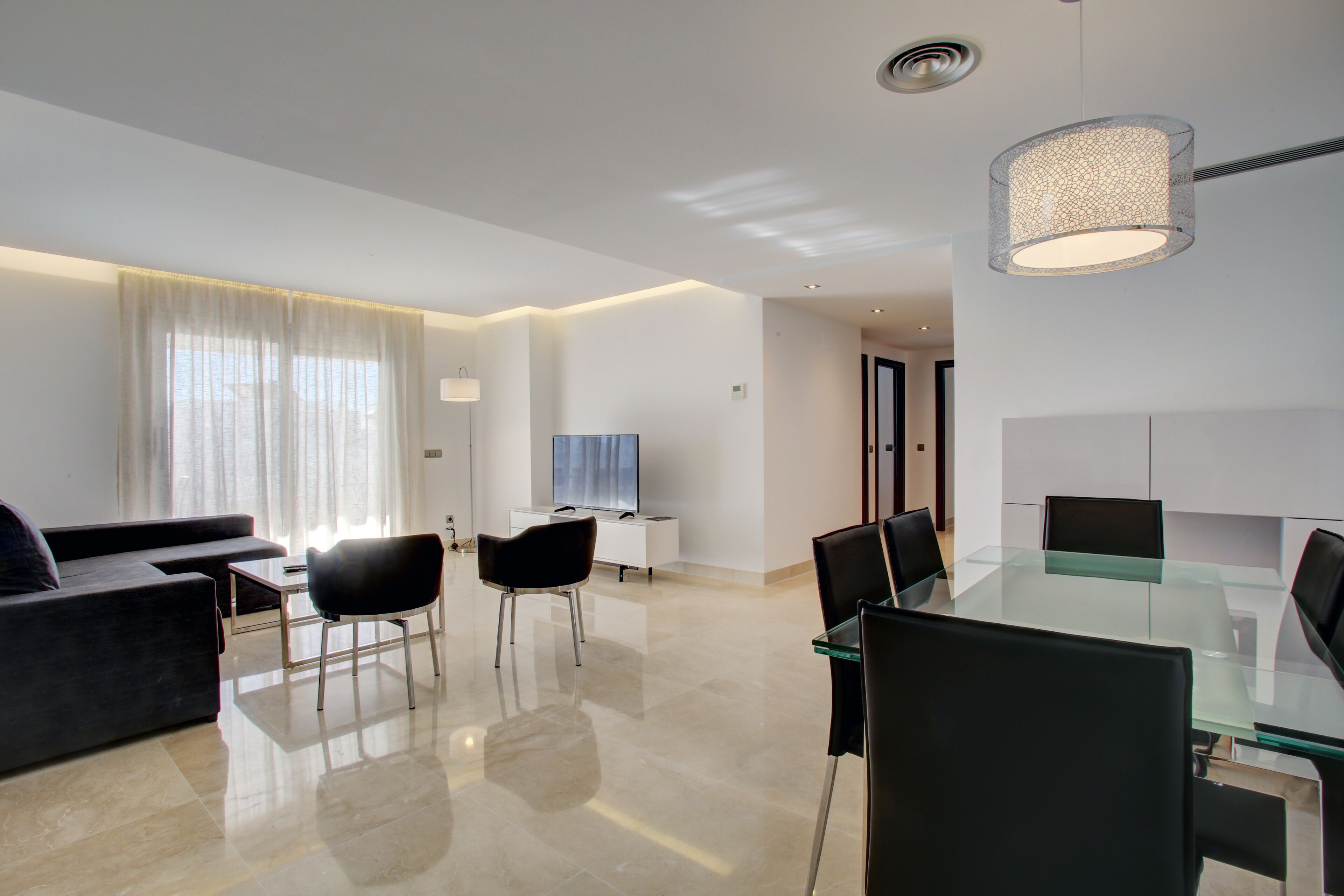 A modern living room with a glass dining table, black chairs, a dark sofa, and a TV.