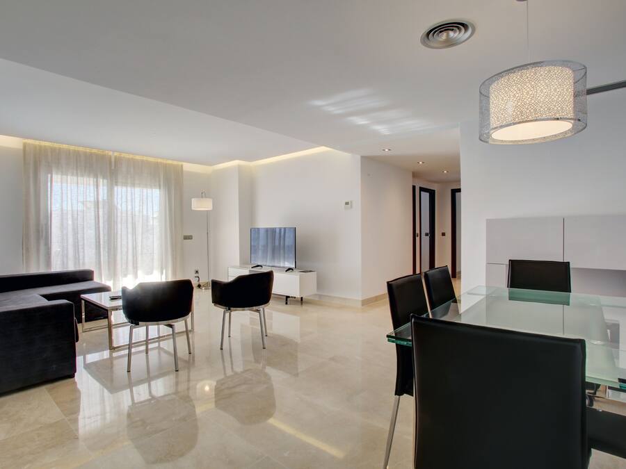 A modern living room with a glass dining table, black chairs, a dark sofa, and a TV.