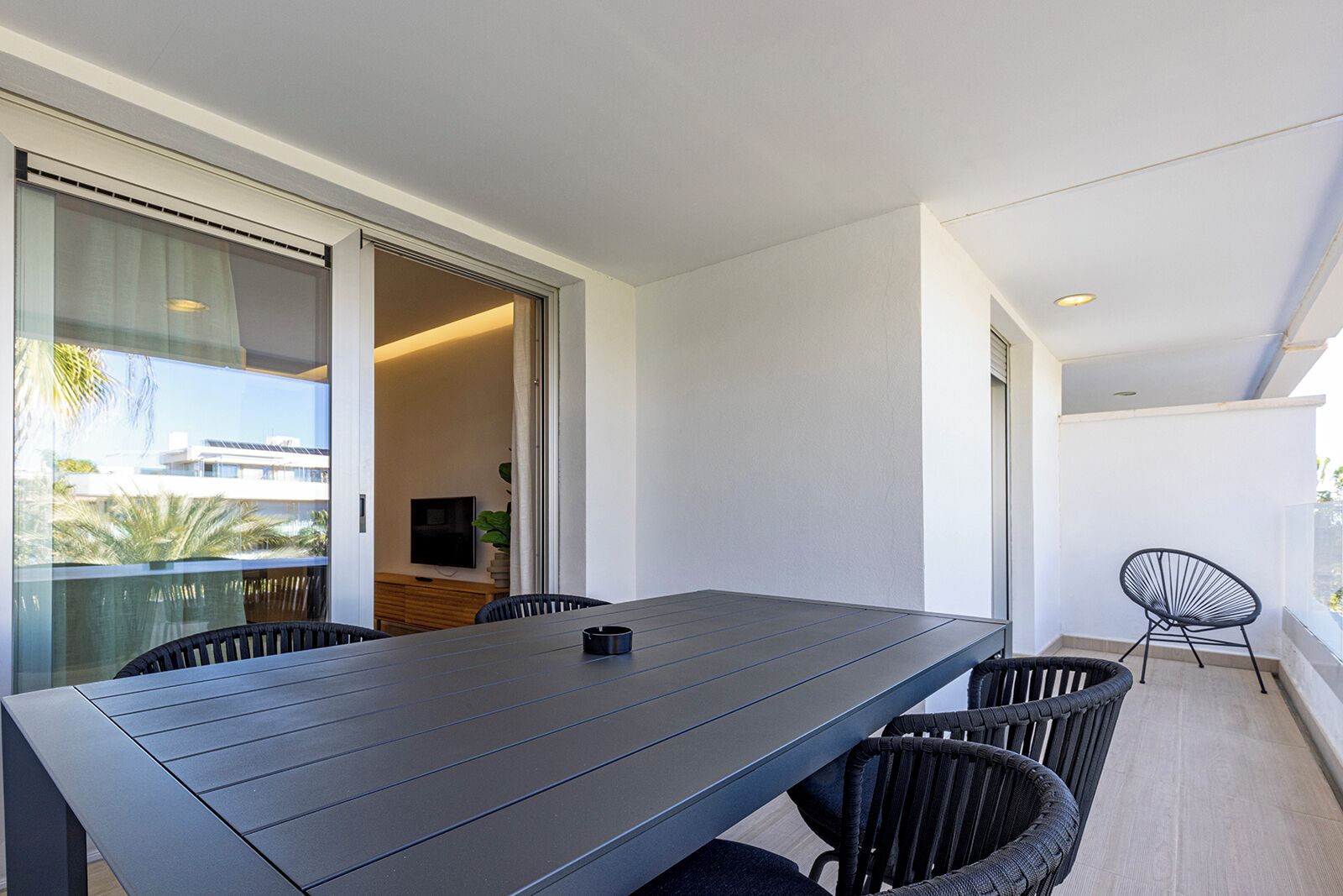 A modern balcony with a dining table and chairs, a TV, and a view of buildings and palm trees.