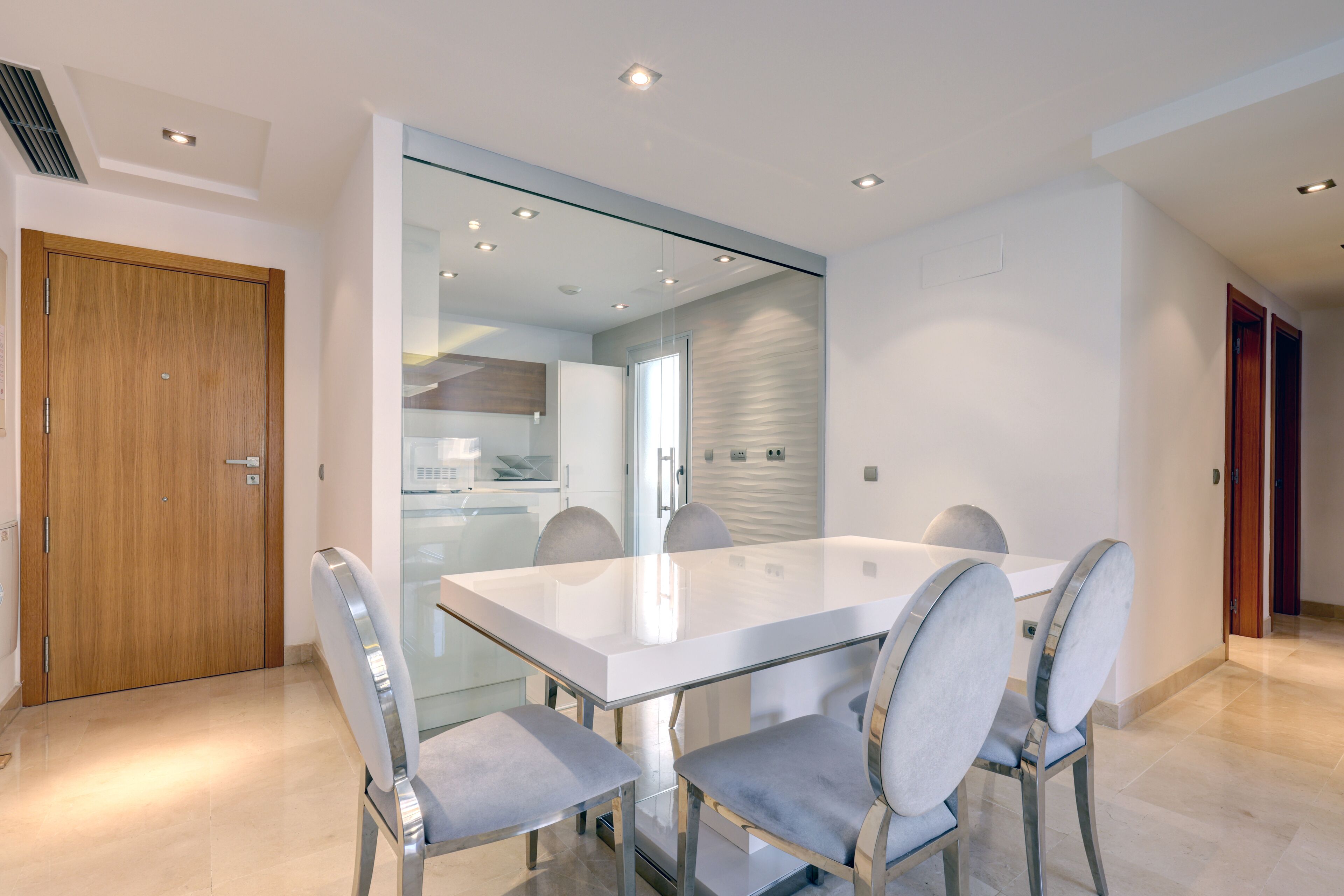 A modern dining area with a glass table and metal chairs, a wooden door, and a kitchen visible through a glass partition.