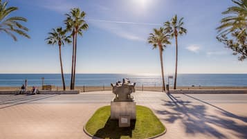 A paved walkway with palm trees and a sculpture, leading to a beach with the ocean in the background.