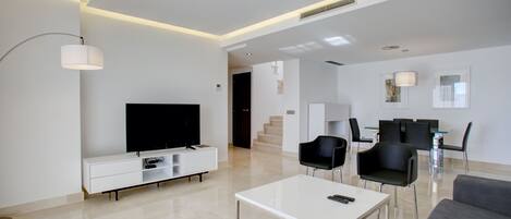 Un salon moderne avec un téléviseur à écran plat, une table basse blanche et des chaises noires. Un escalier mène à un autre niveau.