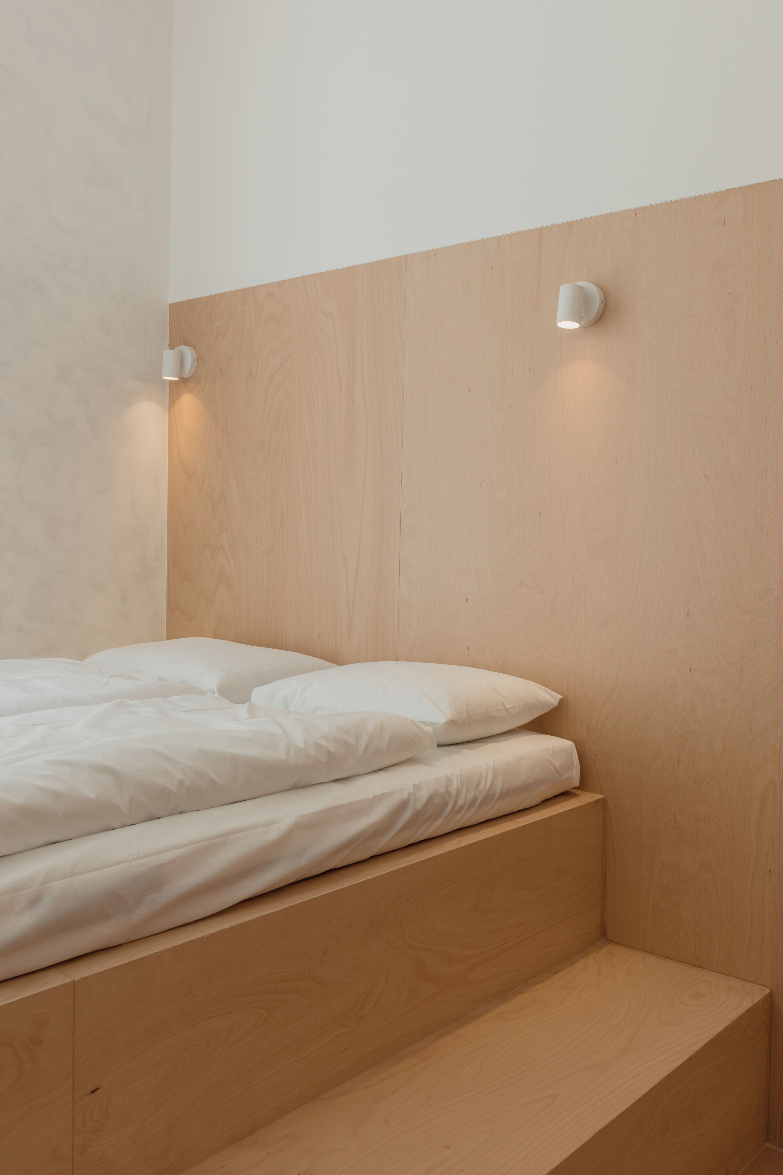 A minimalist bedroom with a wooden headboard, a single bed with white bedding, and two wall-mounted light fixtures.