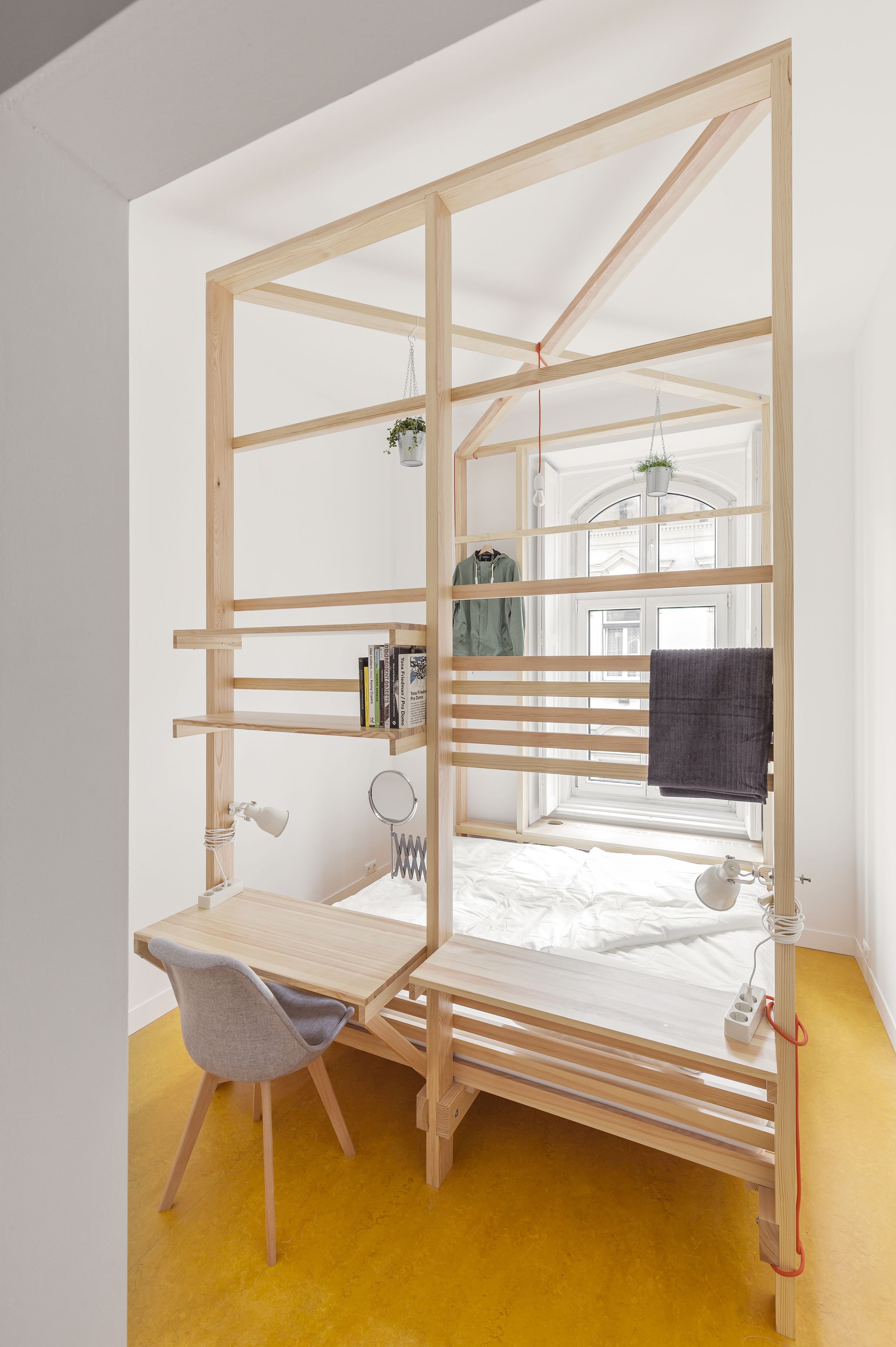 Habitación pequeña con cama, escritorio y silla. Se observan estanterías con libros y una planta colgante.