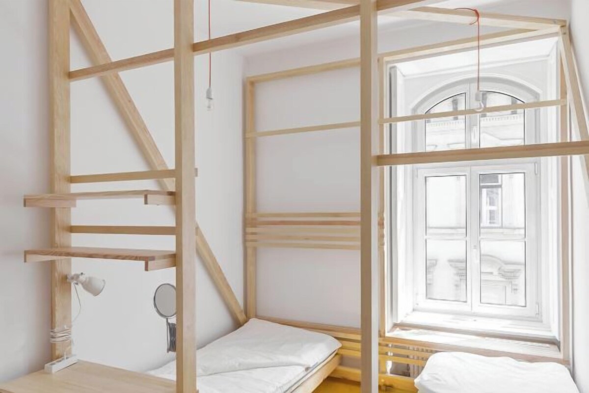 A room with a wooden bunk bed, a small desk with a lamp, and a window with a view of a building.