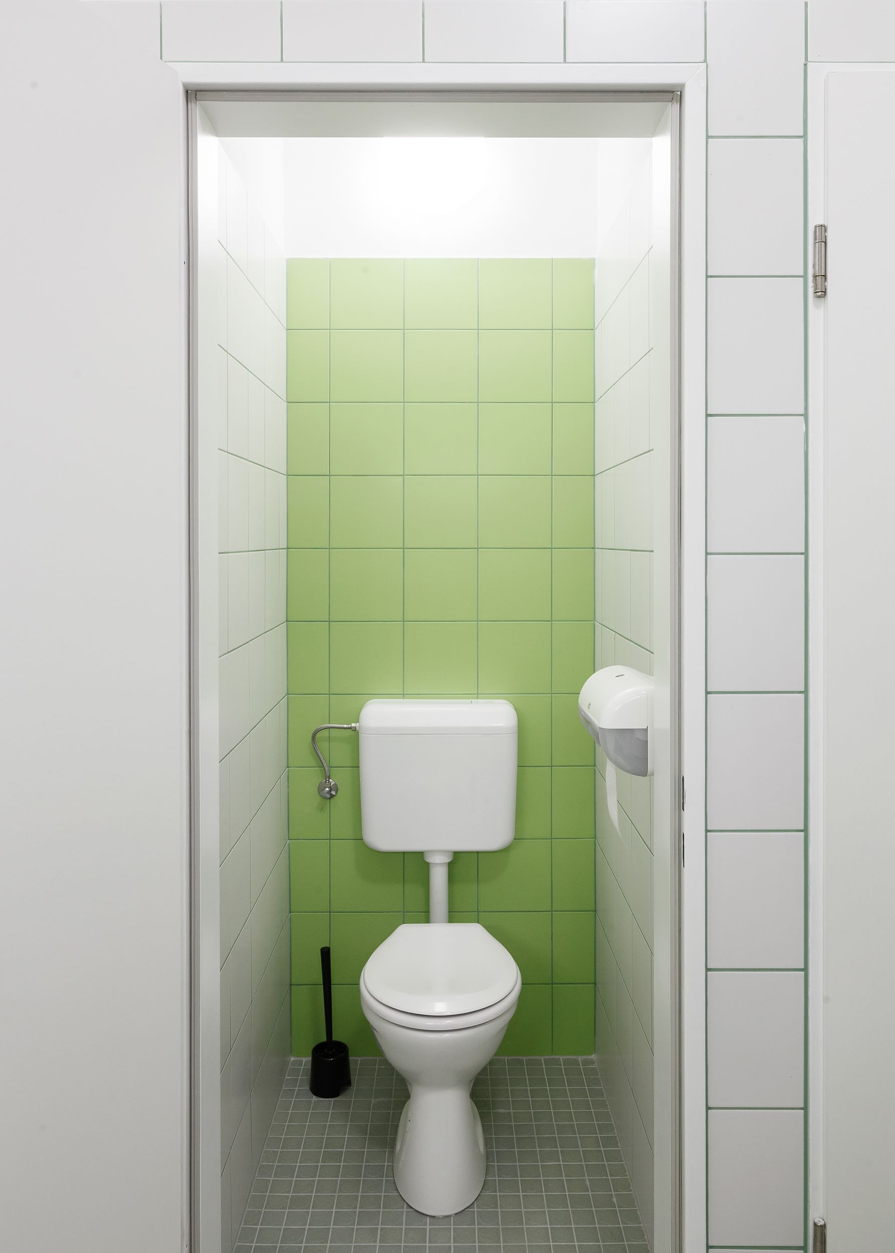 A small, enclosed bathroom with a toilet, a hand dryer, and a plunger.