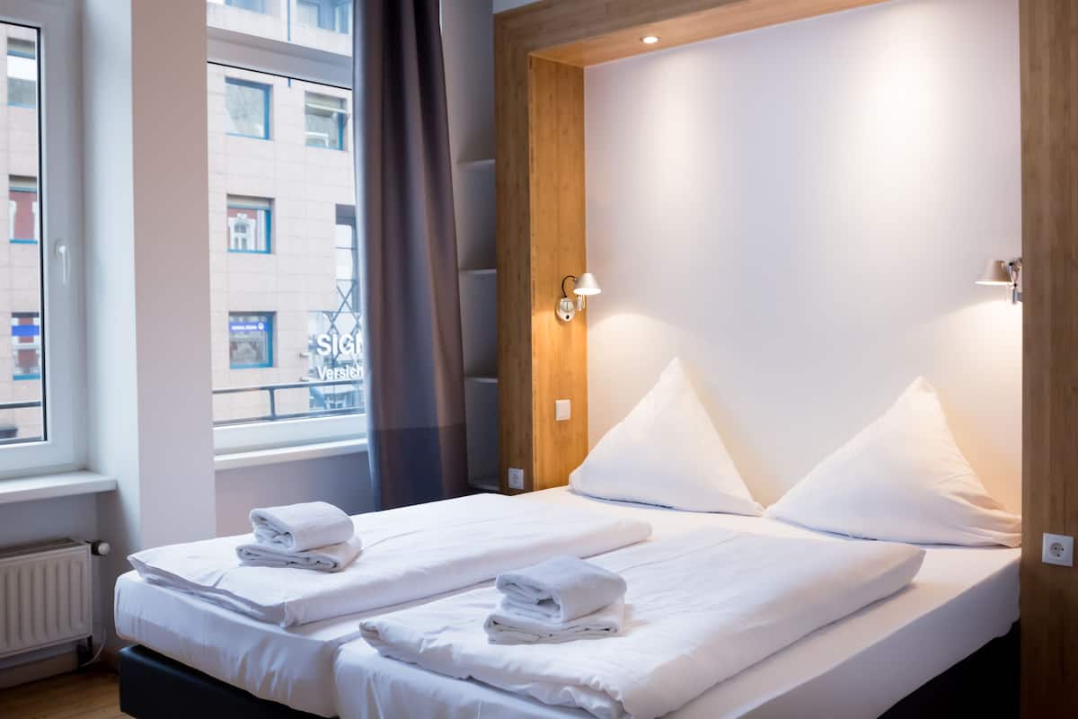 A hotel room with two beds, white linens, a window with a view of buildings, and a wooden accent wall.