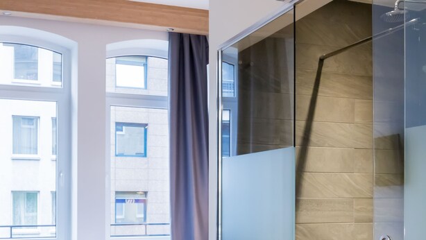 A modern hotel room with a bed, a glass shower enclosure, and large windows.