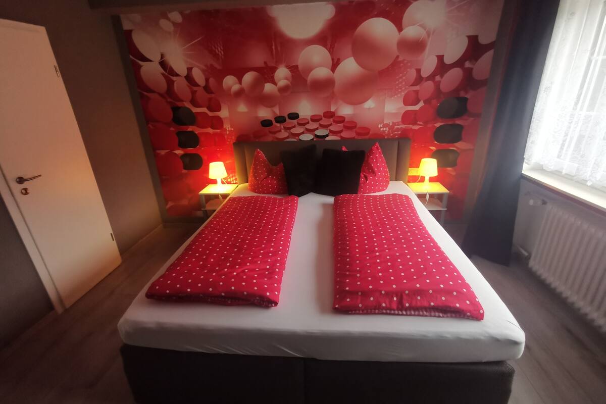 A double bed with red polka dot bedding, two bedside lamps, and a wall with a red and white abstract pattern.
