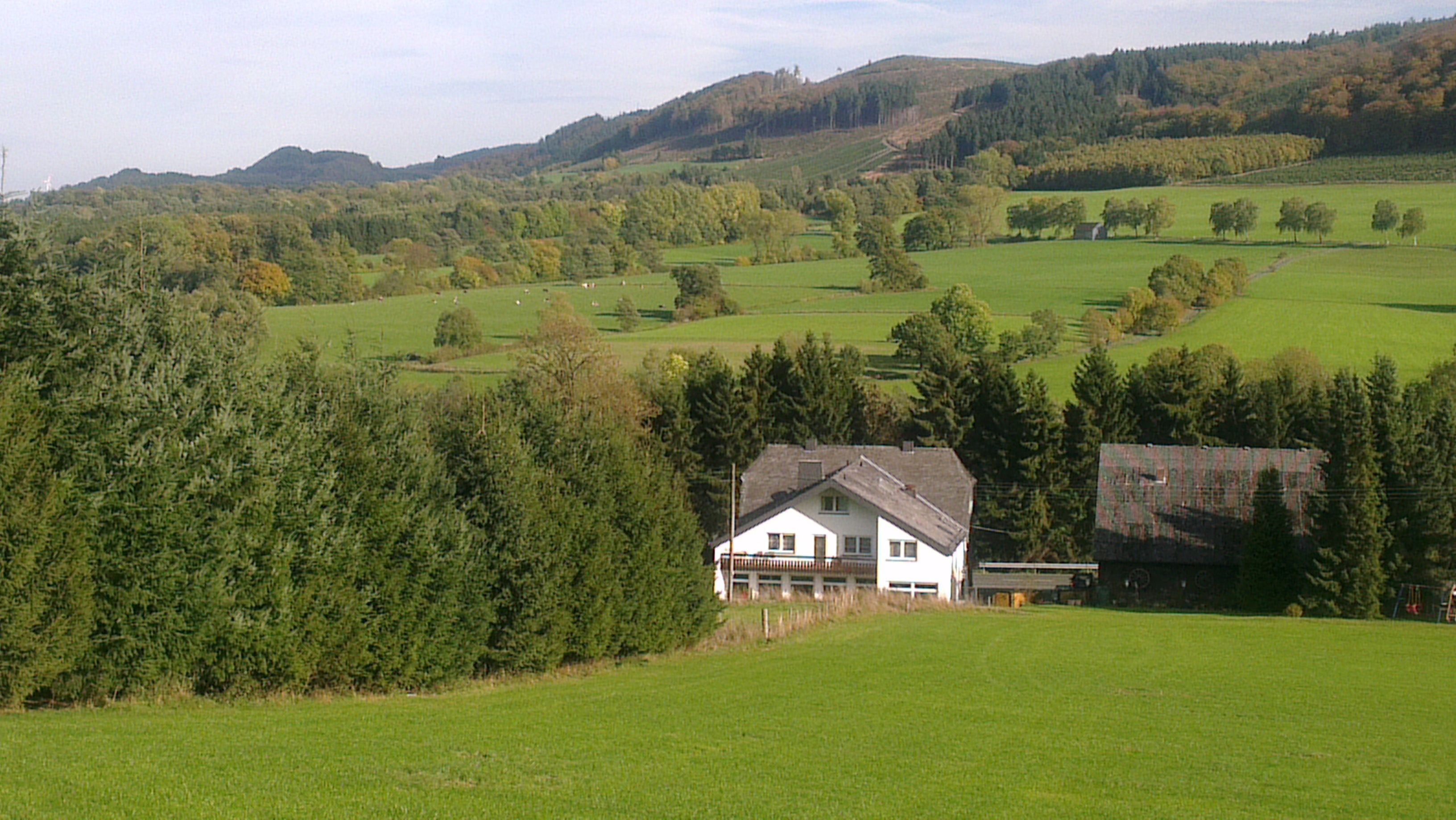 Una casa blanca con balcón, rodeada de campos verdes y árboles, con colinas al fondo.