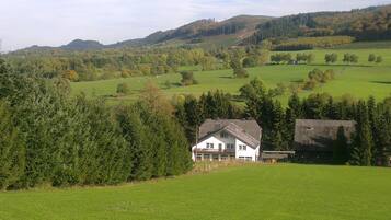 Una casa blanca con balcón, rodeada de campos verdes y árboles, con colinas al fondo.