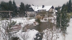 Paisaje nevado con un estanque, un banco de madera y una casa grande con tejado cubierto de nieve.