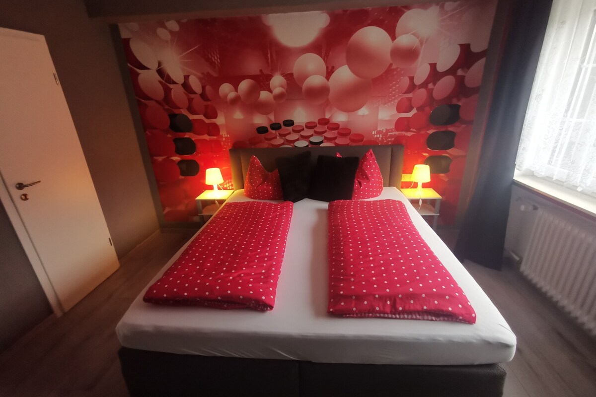 A double bed with red polka dot bedding, two bedside lamps, and a wall with a red and white pattern.