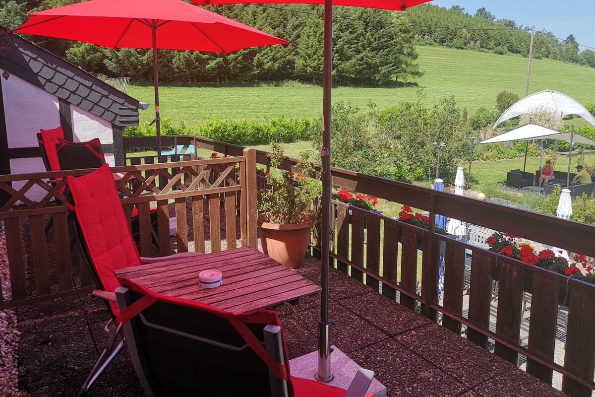A patio with a wooden table and chairs, red umbrellas, and a view of a green field.