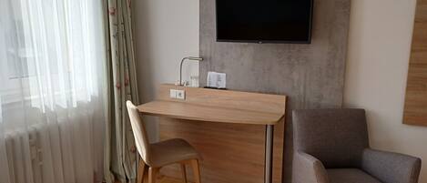 A modern hotel room with a wooden desk, a grey armchair, and a flat-screen TV mounted on the wall.