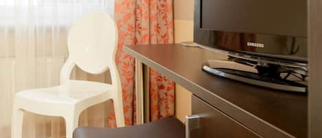 Una habitación de hotel con una televisión sobre un mueble de madera, una silla blanca y un reposapiés marrón.