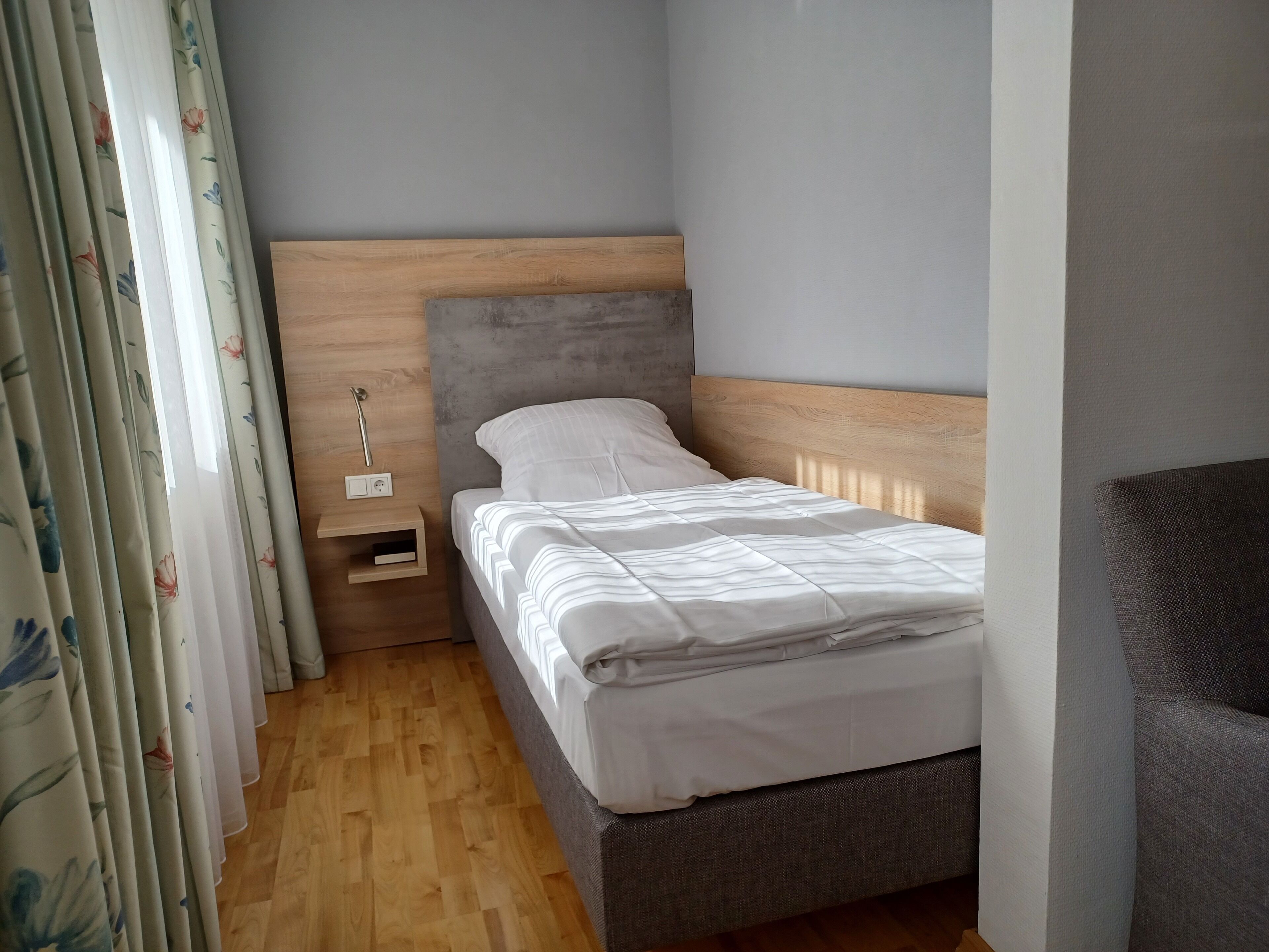 A small, neatly arranged bedroom with a wooden headboard, a single bed, a nightstand, and a wall-mounted power outlet.