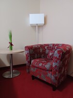A corner room with a red carpet, a floral-patterned armchair, a small round table, and a white floor lamp.