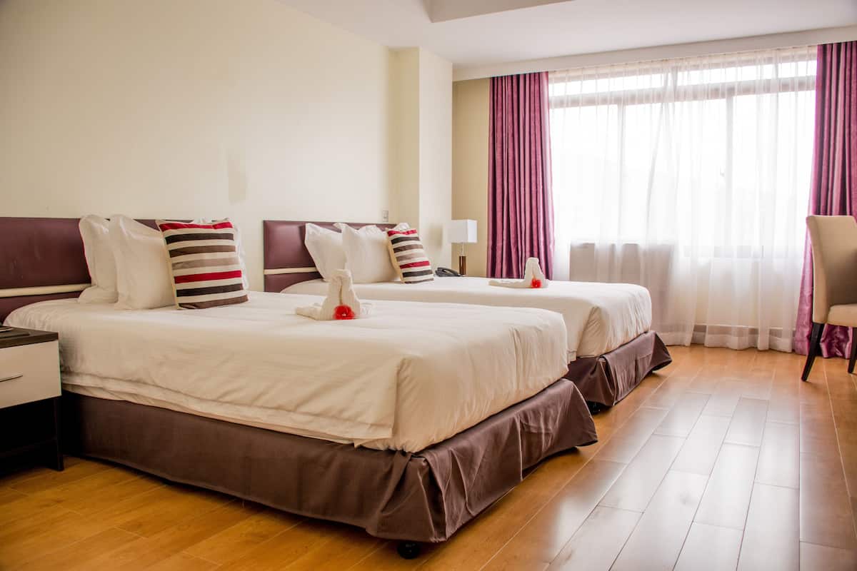A hotel room with two beds, a wooden floor, a nightstand, a lamp, and a chair.