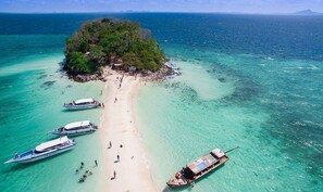 Ein Strand mit klarem türkisblauem Wasser, eine kleine Insel mit dichter Vegetation und mehrere Boote, die in der Nähe des Ufers vor Anker liegen.