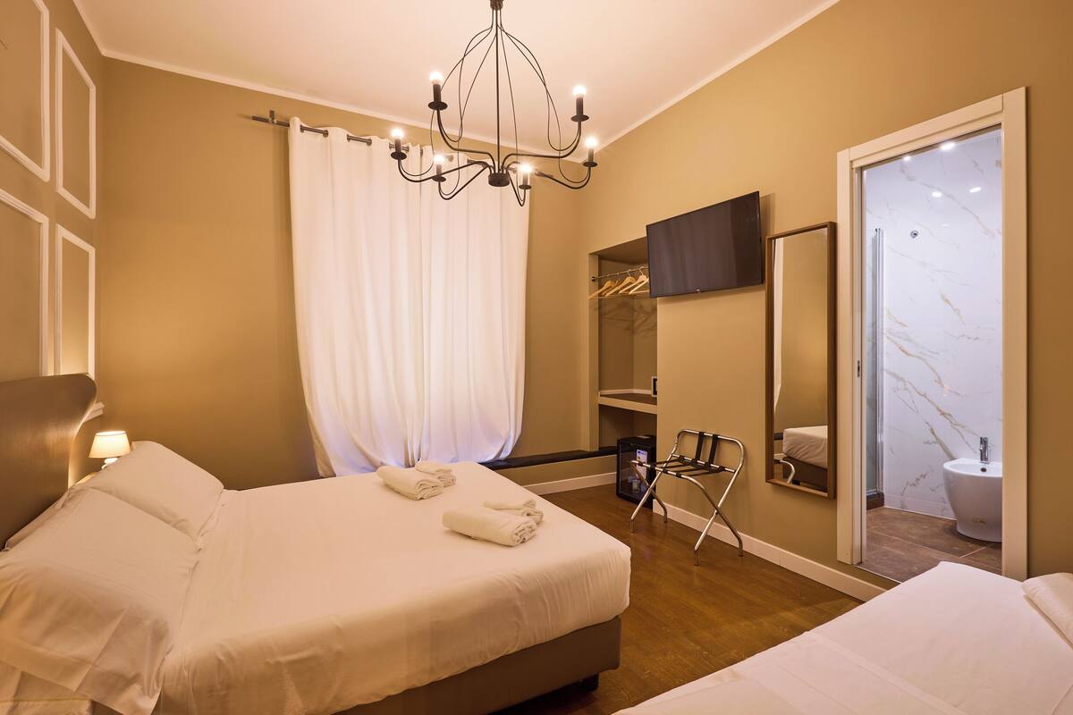 Une chambre d’hôtel avec deux lits, un lustre, un miroir, une télévision et une salle de bain avec un lavabo en marbre.