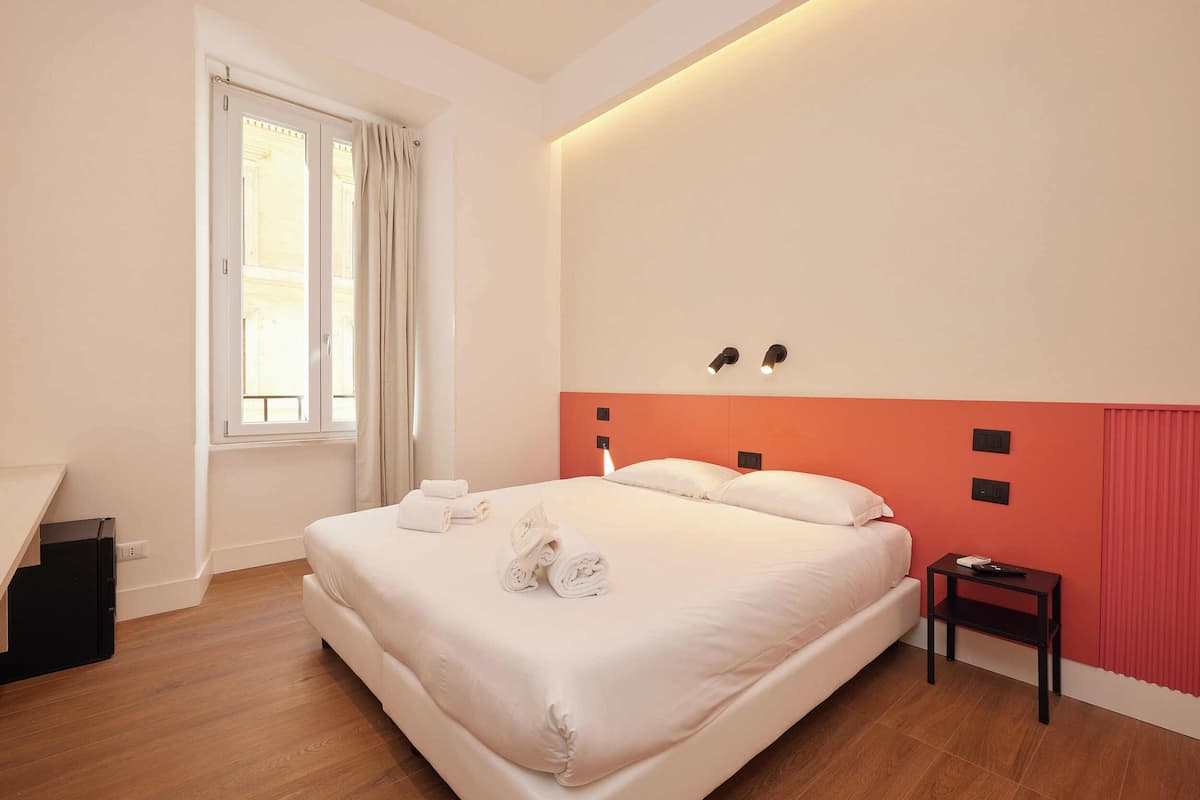 Une chambre d’hôtel avec un lit, une table de chevet, une fenêtre avec des rideaux et un luminaire fixé au mur.