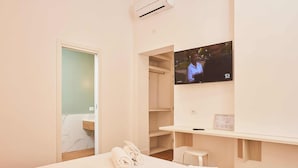 Une chambre d’hôtel avec un lit, un téléviseur fixé au mur, un petit tabouret et une étagère en bois sur laquelle se trouve une télécommande.