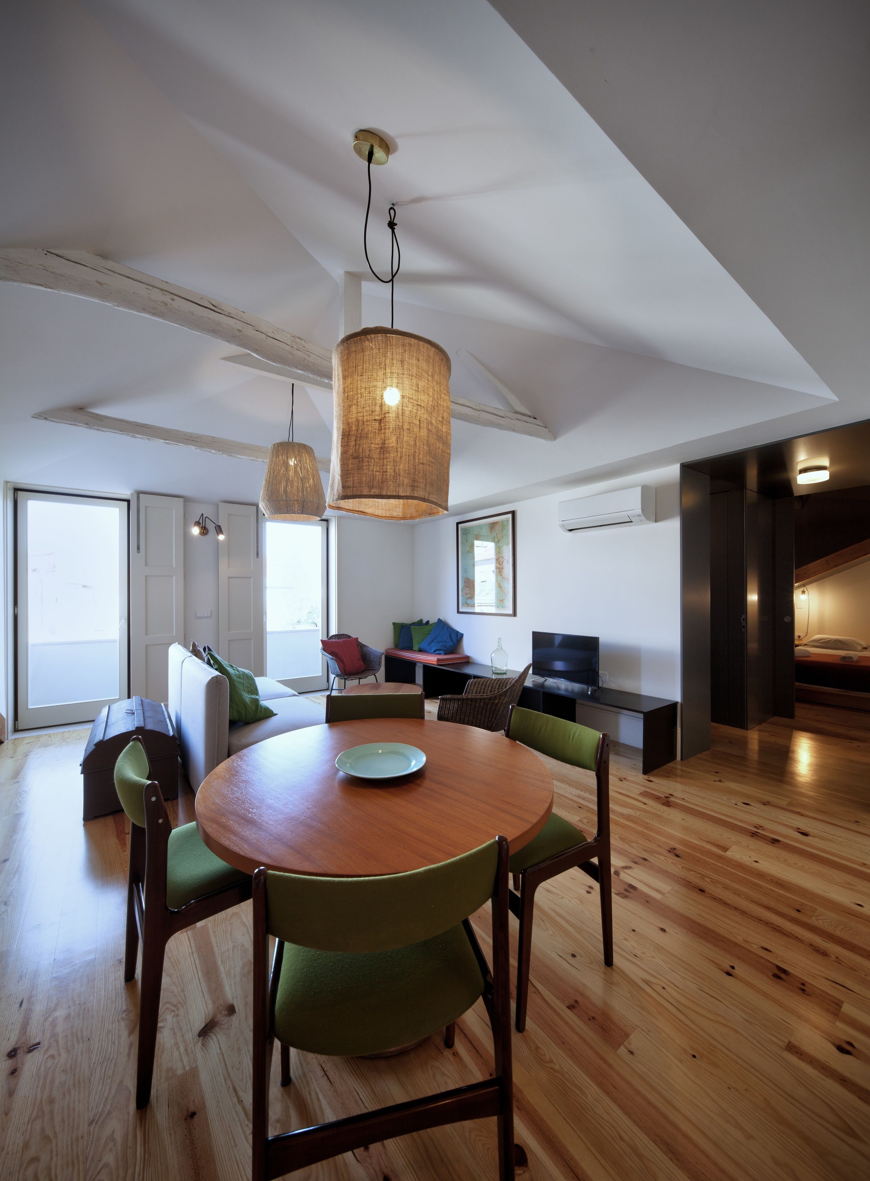 A modern living room with a wooden dining table, green upholstered chairs, a large woven pendant light, and a built-in TV.