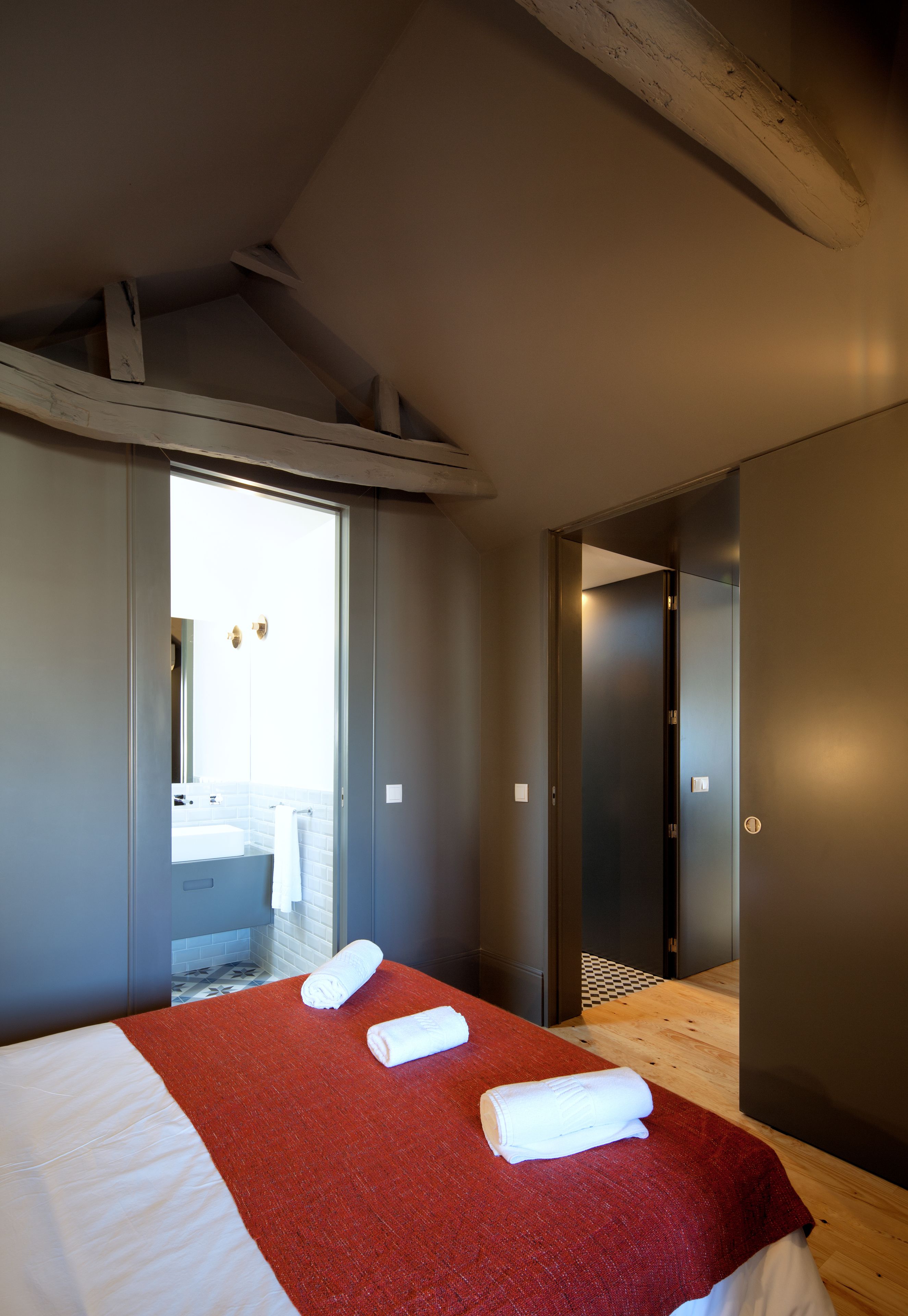 Una habitación de hotel moderna con una cama grande, una colcha roja y dos toallas enrolladas. Se ve un baño a través de una puerta de cristal, y el suelo es de madera.